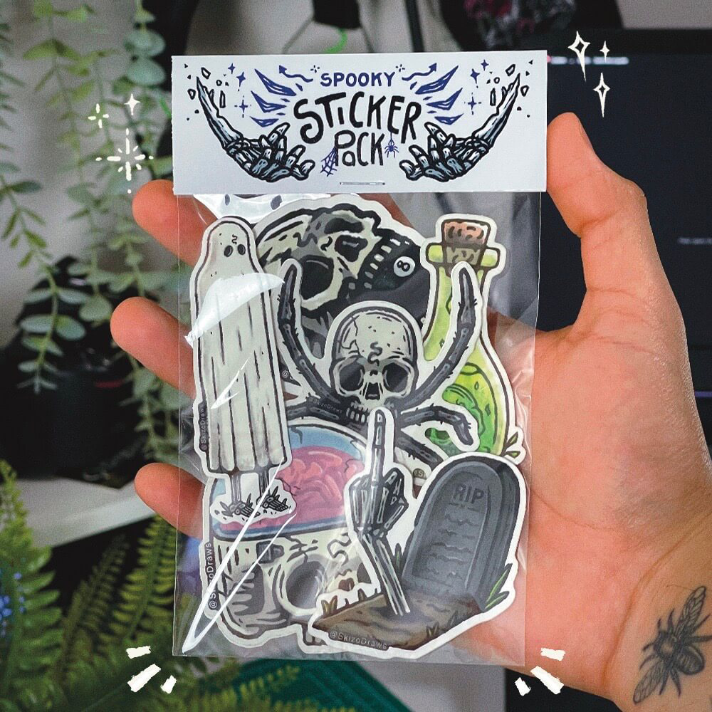 En hånd holder en klistermærke-pakke med uhyggelige tegneseriestickers og en header, hvor der står "SPOOKY STICKER PACK", foran en baggrund af planter