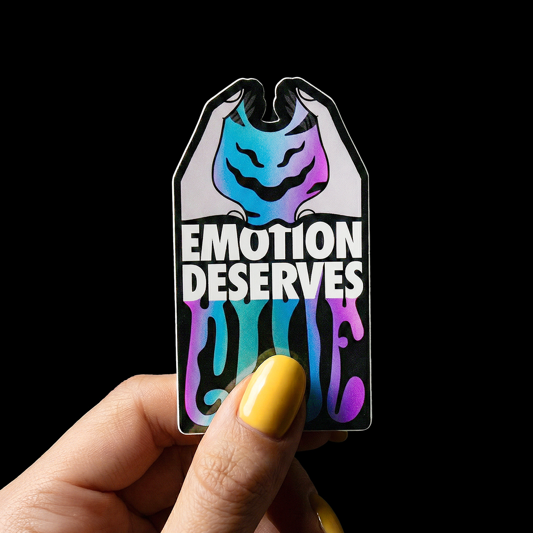A hand with yellow nails holds a colorful sticker reading "Emotion Deserves Love" with a smiling blue and purple face.