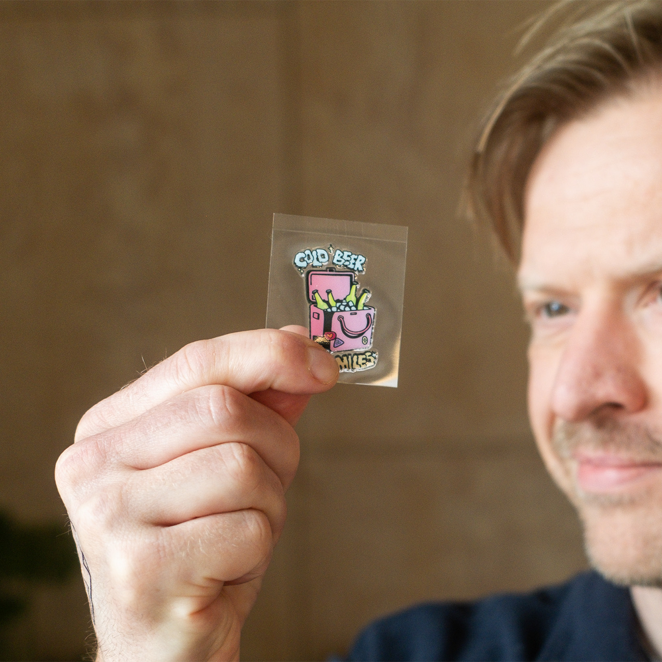 Person holding a small transparent sticker with a colorful illustration of a pink container with fries and the text “Cold beer, long miles.”
