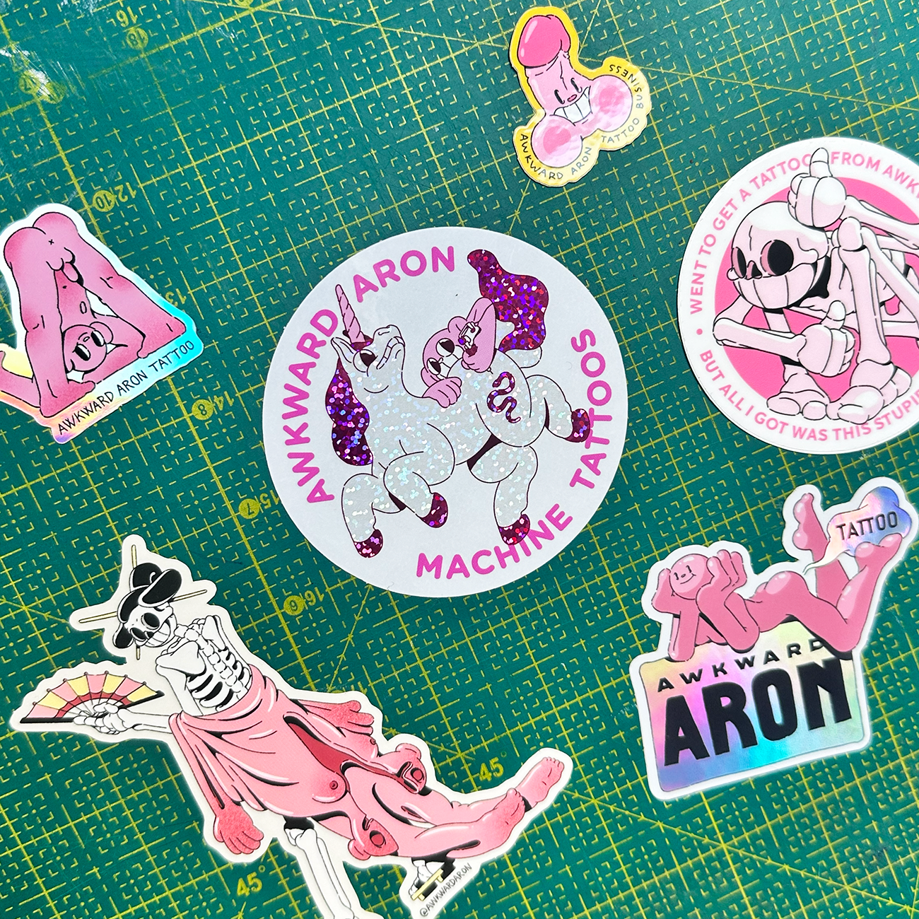 Die-cut stickers from tattoo artist Awkward Aron, printed on glitter, holographic and vinyl, set on a cutting mat background.