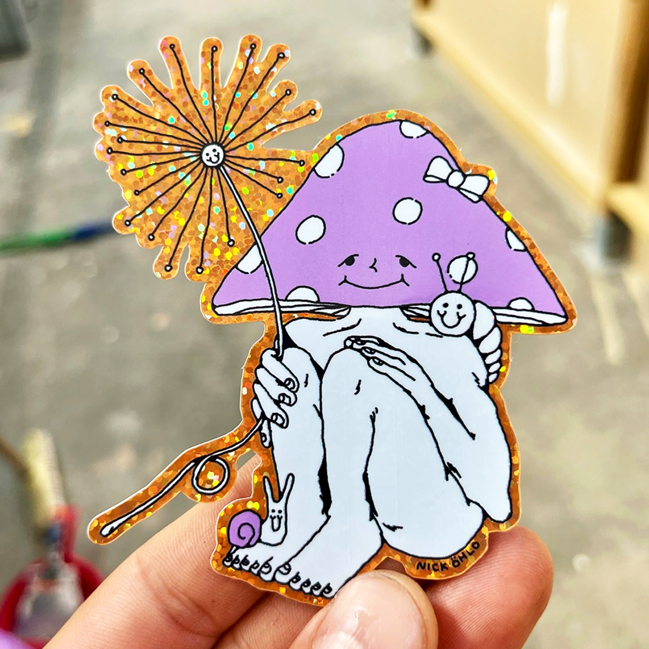 Hand holding a glitter sticker of a mushroom man with a flower as parasol, against a blurry background.