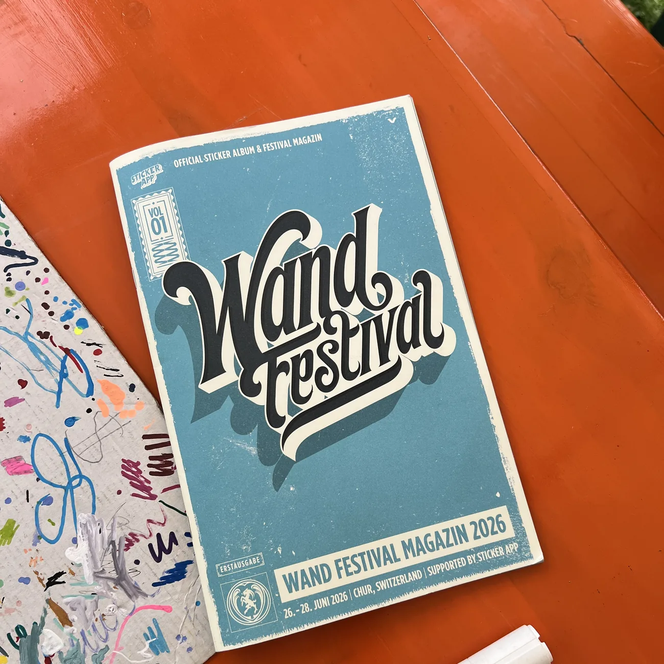 Wand Festivals officielle klistermærkealbum og festivalmagasin 2026, støttet af StickerApp, ved siden af en malertusch på et orange bord
