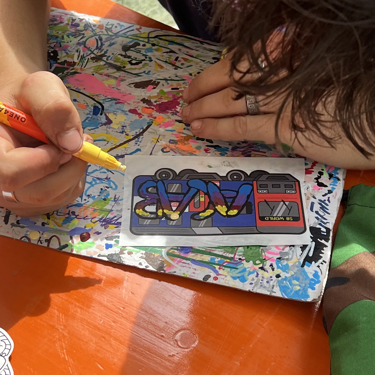 En hånd farvelægger en etiket med S8 World-toget med en gul malertusch på workshoppen med Brainfart x StickerApp