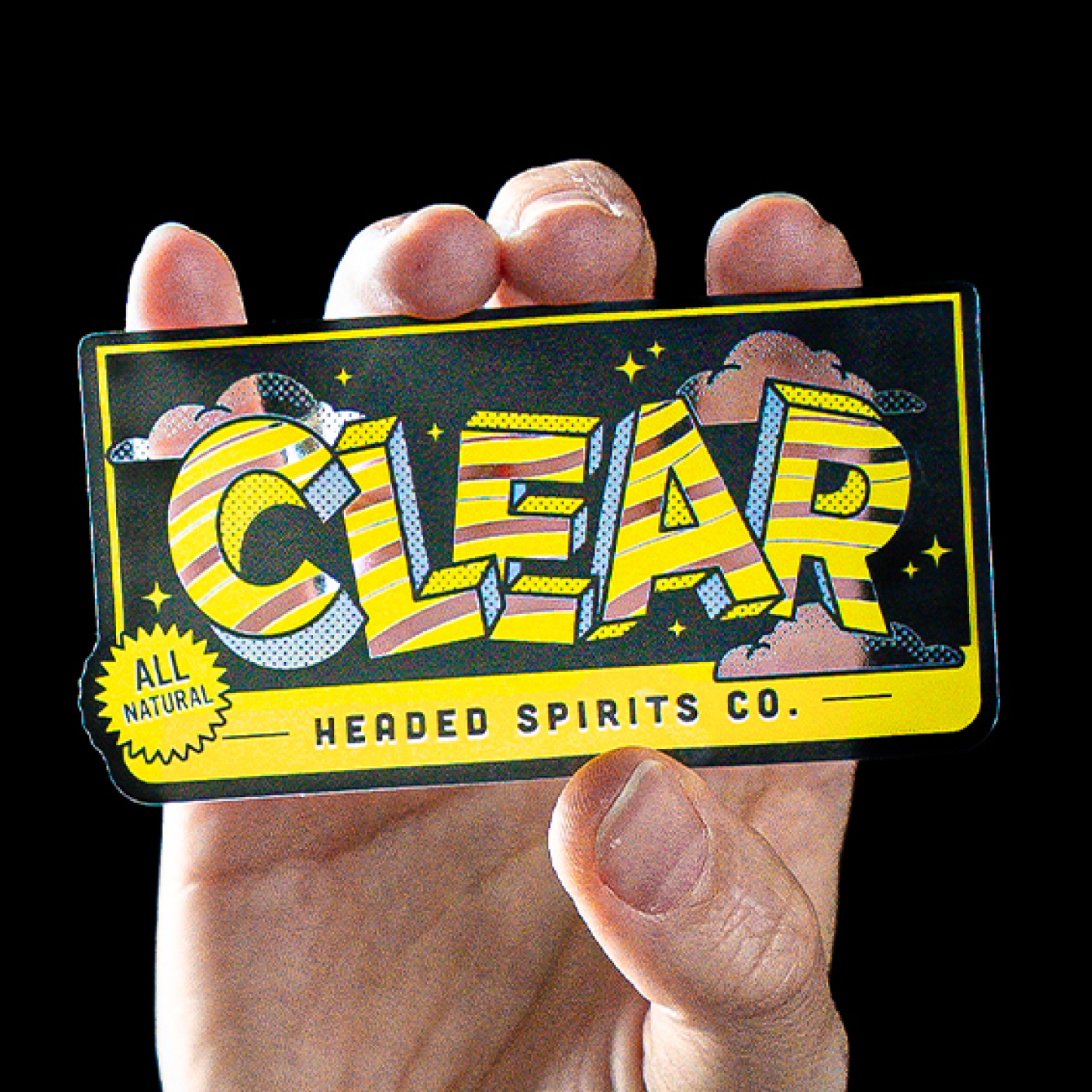 Gennemsigtigt klistermærke med gult og sort "Clear Headed Spirits Co."-design, holdt i en hånd