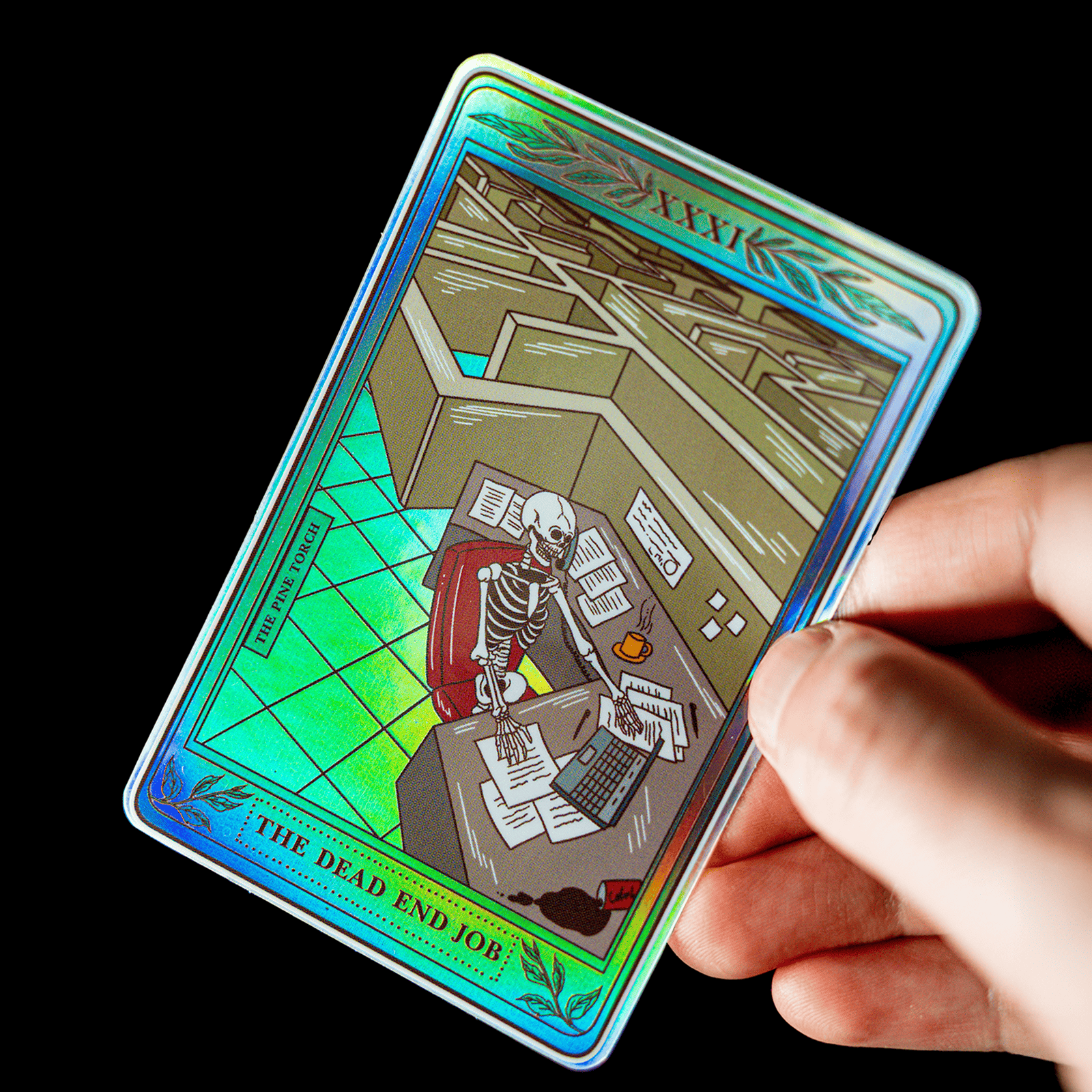 Hånd, der holder et holografisk klistermærke formet som et tarotkort med titlen ”The Dead End Job” og et skelet ved et kontorskrivebord
