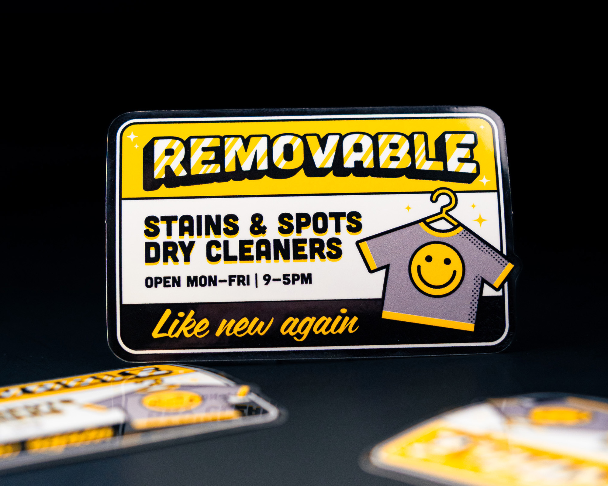 A rectangular sticker for "Removable Stains & Spots Dry Cleaners" featuring a shirt with a smiley face, hours listed as Mon-Fri, 9-5pm.