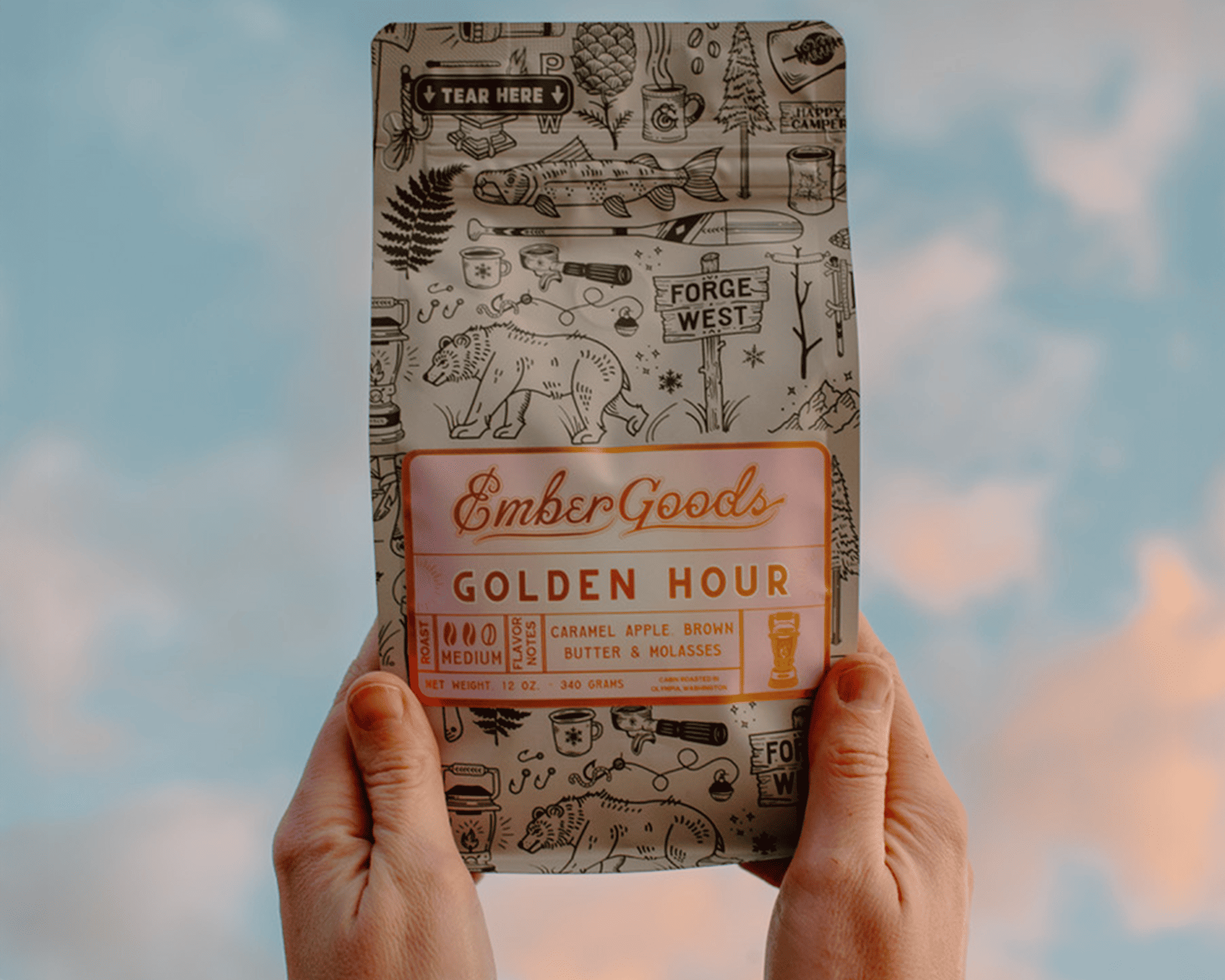 Hænder, der holder en pose Ember Goods Golden Hour-kaffe med en metallisk etiket mod en himmel i baggrunden