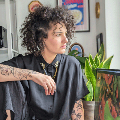 Tattooed Woman looking out a window with a computer screen and plant in the foreground.