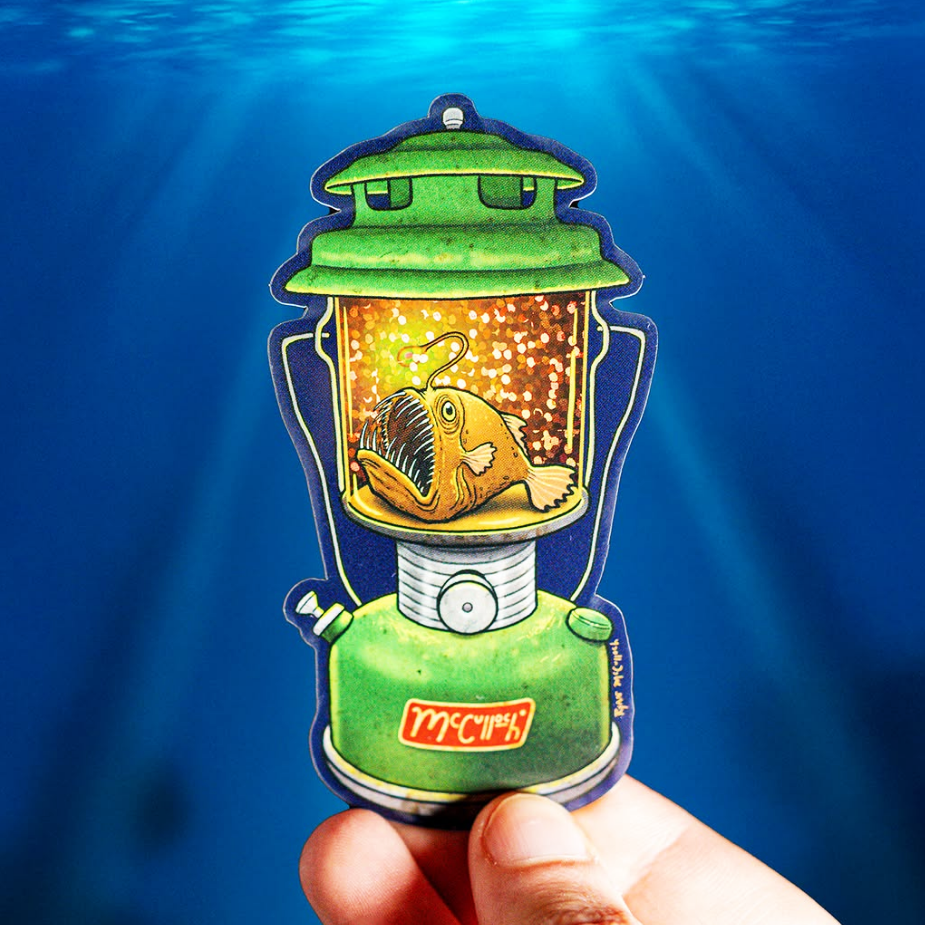 Die-cut glitter sticker with an angler fish stuck in a green oil lamp in a deep blur ocean background